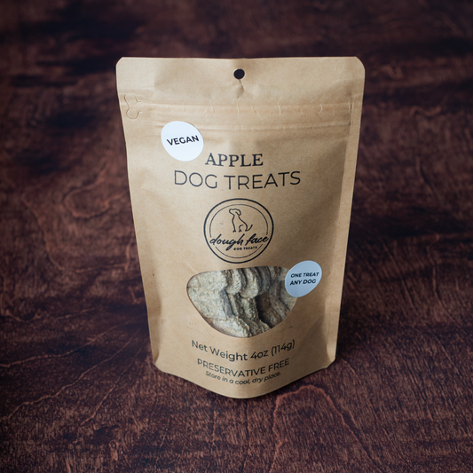 Brown kraft paper stand up pouch of dog treats on a brown wooden background. The words APPLE DOG TREATS are above a logo and a window showing the dog treats inside. There is a white sticker in the top left hand corner of the package with the word VEGAN in black letters.