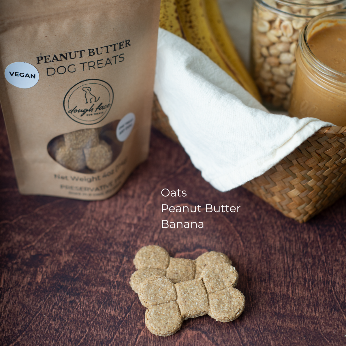 A brown kraft paper stand-up pouch labeled 'PEANUT BUTTER DOG TREATS' with a white sticker that reads VEGAN sits on a dark wooden surface beside a woven basket holding ripe bananas, a mason jar of whole peanuts, and a mason jar of peanut butter. There are two bone-shaped dog treats scattered in the foreground. The pouch appears to be the product being sold, while the basket and food props suggest flavor or ingredients. The ingredients are listed: Oats, Peanut Butter, Banana.