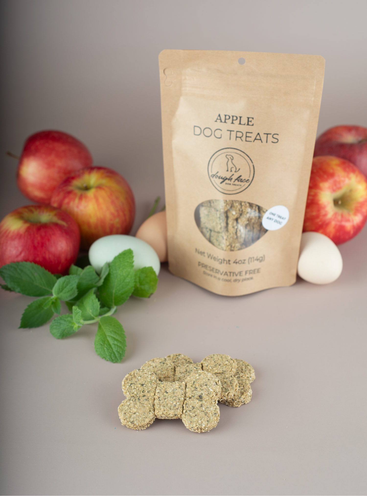 A stand-up kraft paper pouch labeled 'APPLE DOG TREATS' with a clear window showing the treats inside, styled on a neutral surface with red apples, mint leaves, and two eggs around it. A few bone-shaped dog treats are placed in front of the pouch.