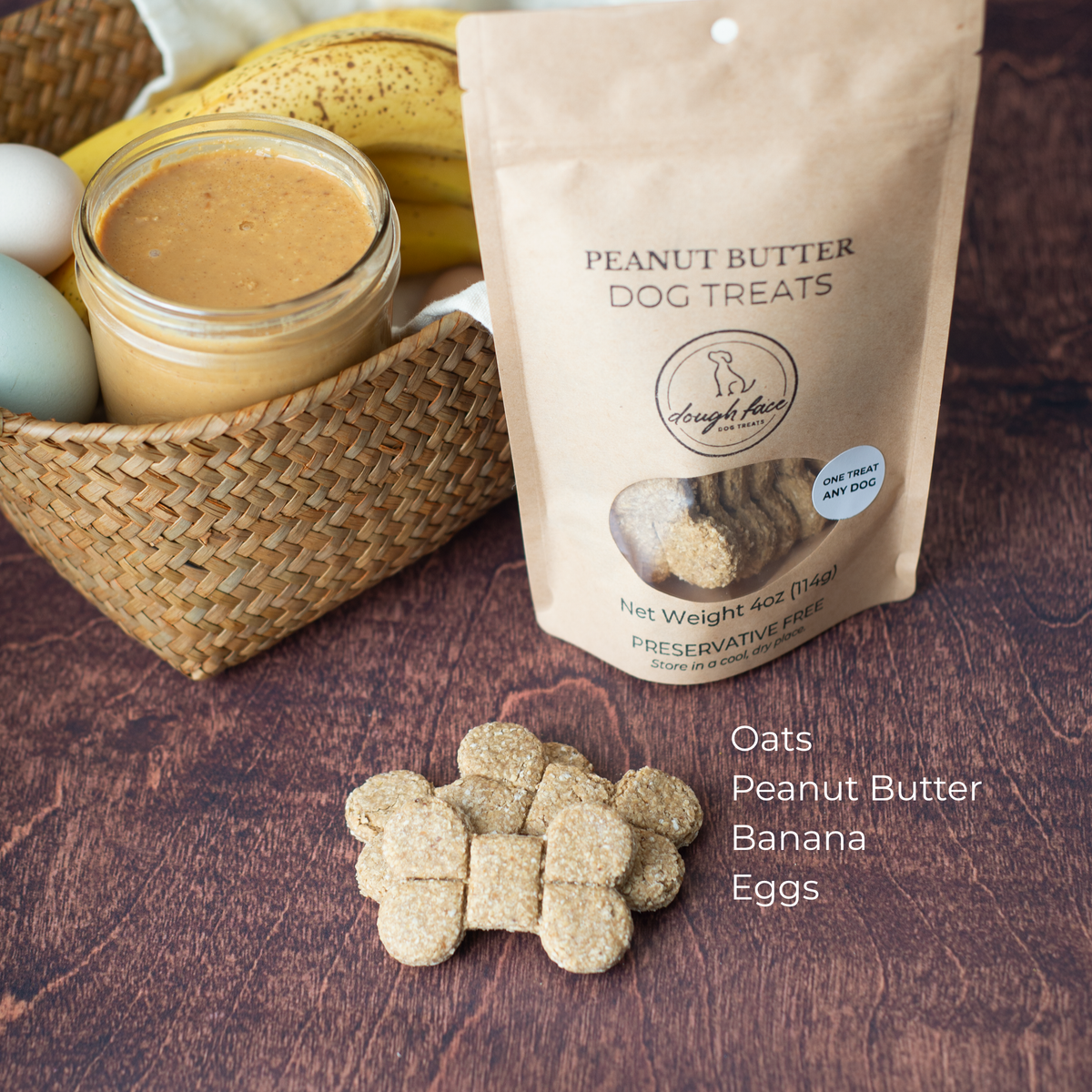 A stand-up pouch labeled 'PEANUT BUTTER DOG TREATS' sits on a dark wooden surface beside a woven basket holding bananas, a jar of peanut butter, and eggs. A few dog treats are scattered in front. The pouch appears to be the product being sold, while the basket and food props suggest flavor or ingredients.