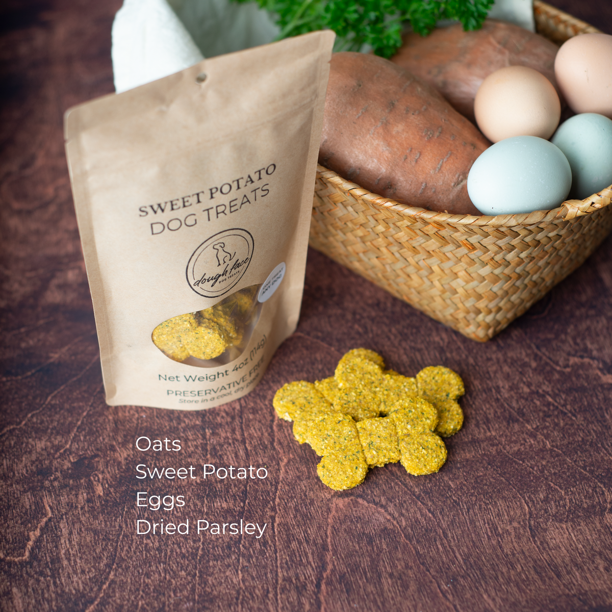 A brown kraft paper stand-up pouch labeled 'SWEET POTATO DOG TREATS' sits on a dark wooden surface beside a woven basket holding two sweet potatoes, four eggs, and fresh parsley. There are three bone-shaped dog treats scattered in the foreground. The pouch appears to be the product being sold, while the basket and food props suggest flavor or ingredients. The ingredients are listed: Oats, Sweet Potato, Eggs, Dried Parsley.