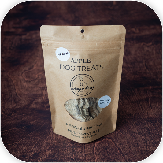 A 4 oz stand-up pouch of Dough Face Dog Treats Apple Dog Treats, displayed on a dark wood background. The packaging is a natural kraft paper style with a clear window showing the treats inside.