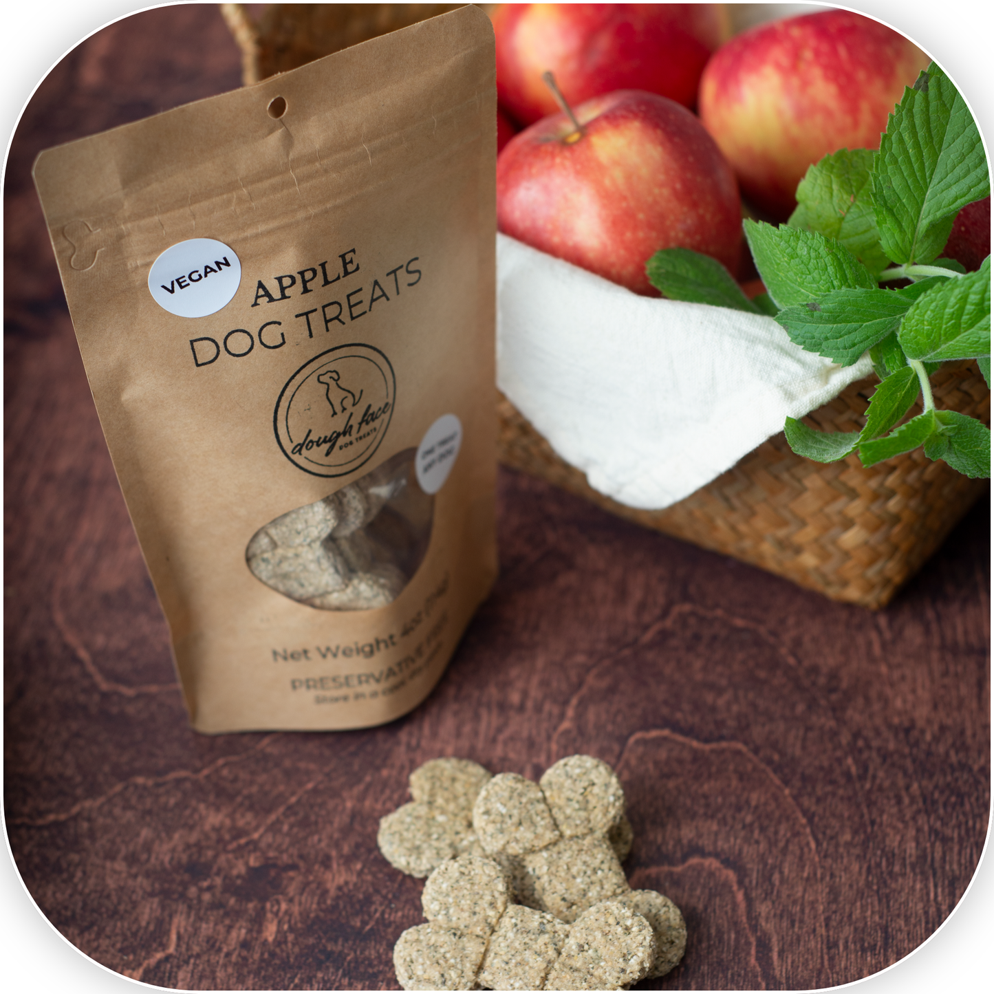 A stand-up pouch of Apple Dog Treats by Dough Face Dog Treats sits on a dark wooden surface next to fresh red apples, mint leaves, and a woven basket. Several bone-shaped treats are placed in front of the bag.