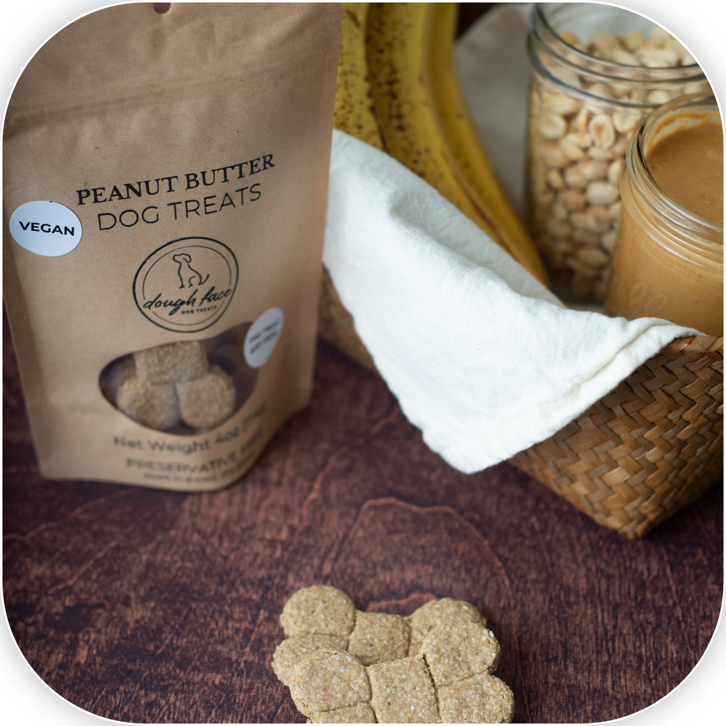 A bag of Dough Face Dog Treats Peanut Butter Dog Treats stands on a dark wooden surface next to a woven basket, white cloth, banana, and jars of peanut butter and peanuts. A peanut butter treat is placed in the foreground.