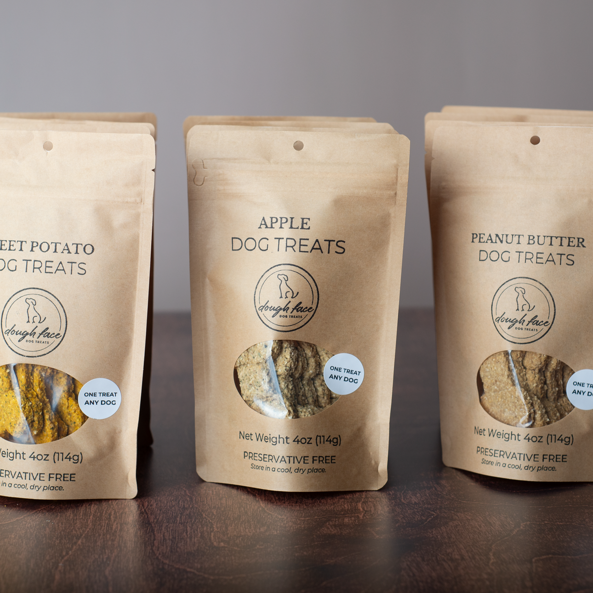 Three stand-up pouches of Dough Face Dog Treats brand dog treats on a dark wooden surface against a gray background. The center pouch is labeled Apple Dog Treats, flanked by Sweet Potato Dog Treats on the left and Peanut Butter Dog Treats on the right.
