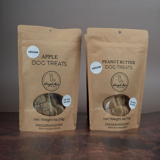 Two brown kraft stand up pouches on a dark wood textured table with a gray background. Each bag has black lettering "Net Weight 4oz (114g) PRESERVATIVE FREE Store in a cool dry place". There is a window in each bag showing the dog treats inside and a white sticker with the word VEGAN in black text.