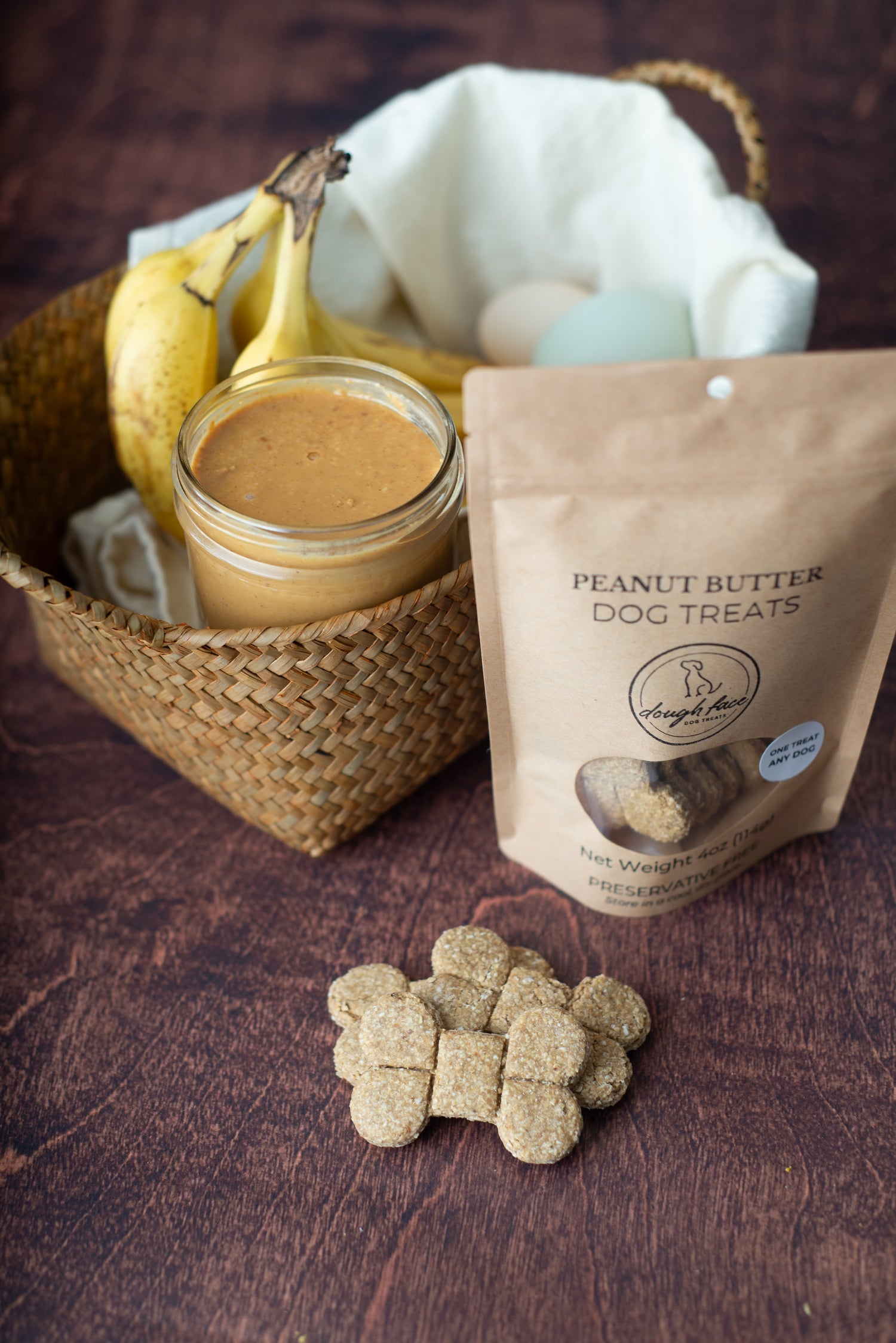 A stand-up pouch of Dough Face Dog Treats brand 'Peanut Butter' dog treats stands on a dark wooden surface next to a woven basket containing bananas, a jar of peanut butter, and eggs, with several treats scattered in front.