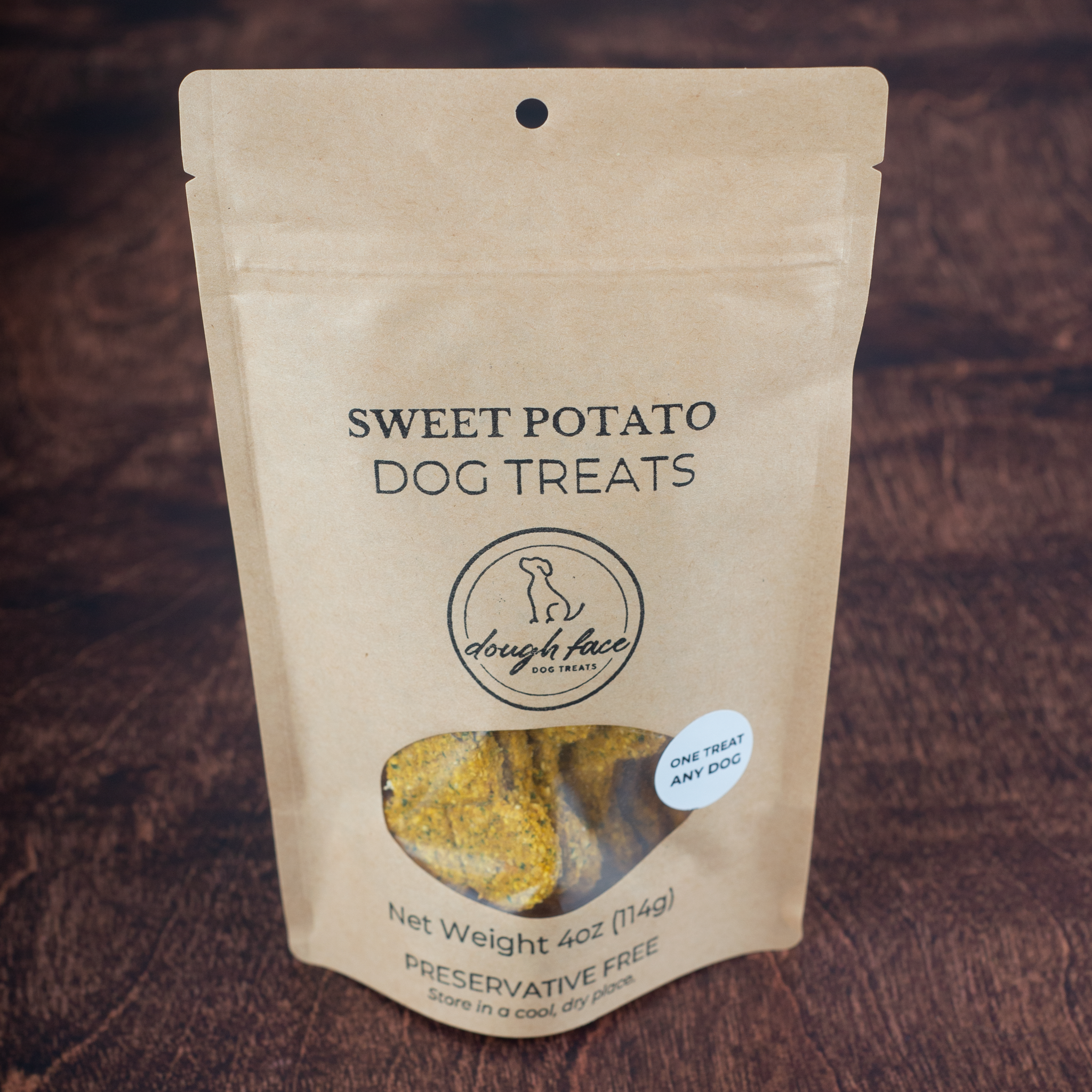 A stand-up kraft paper pouch of dog treats on a dark wood background. The package is labeled 'SWEET POTATO DOG TREATS' with a circular logo reading 'dough face' and a clear window showing treats inside.