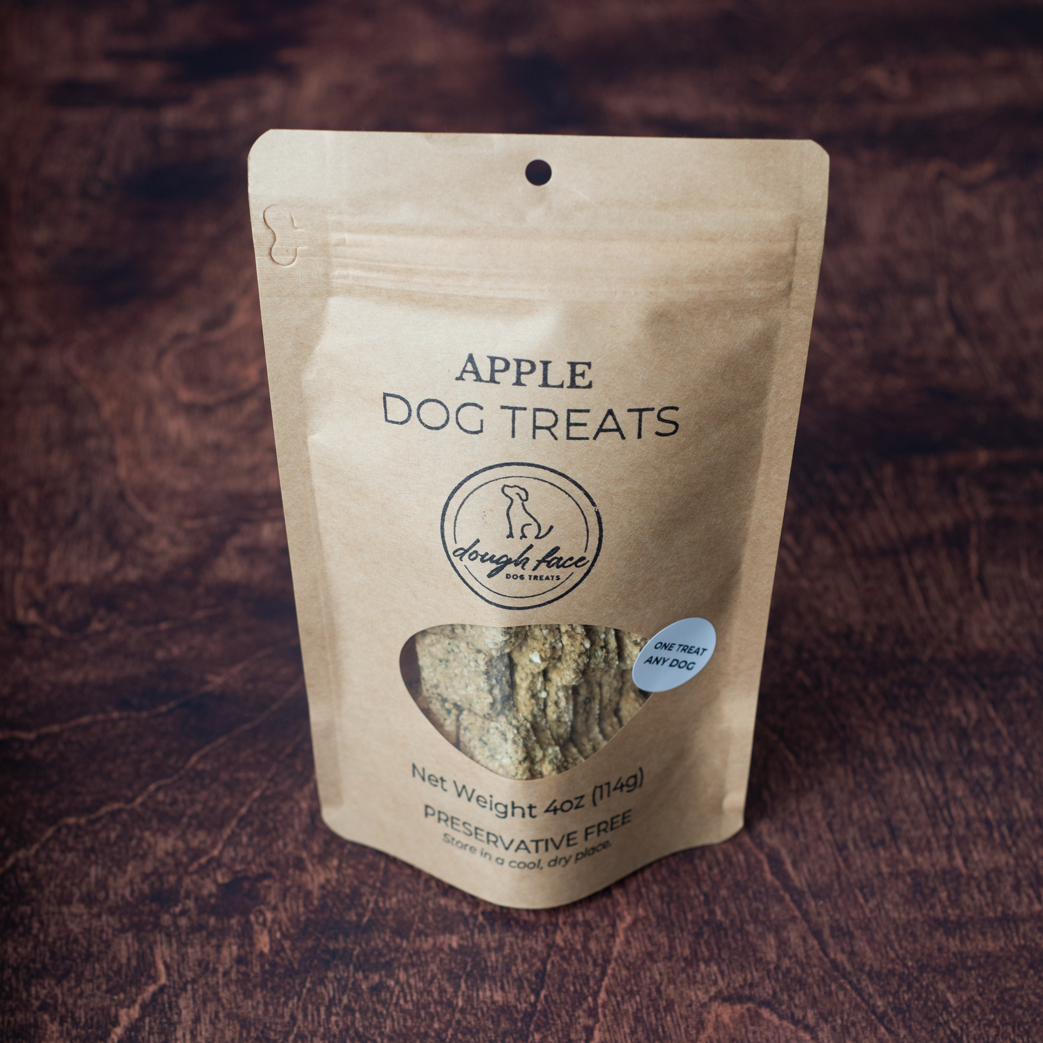 A stand-up pouch of Apple Dog Treats by Dough Face Dog Treats, shown on a dark wood background. The kraft paper pouch features a clear window displaying the apple-flavored treats inside.