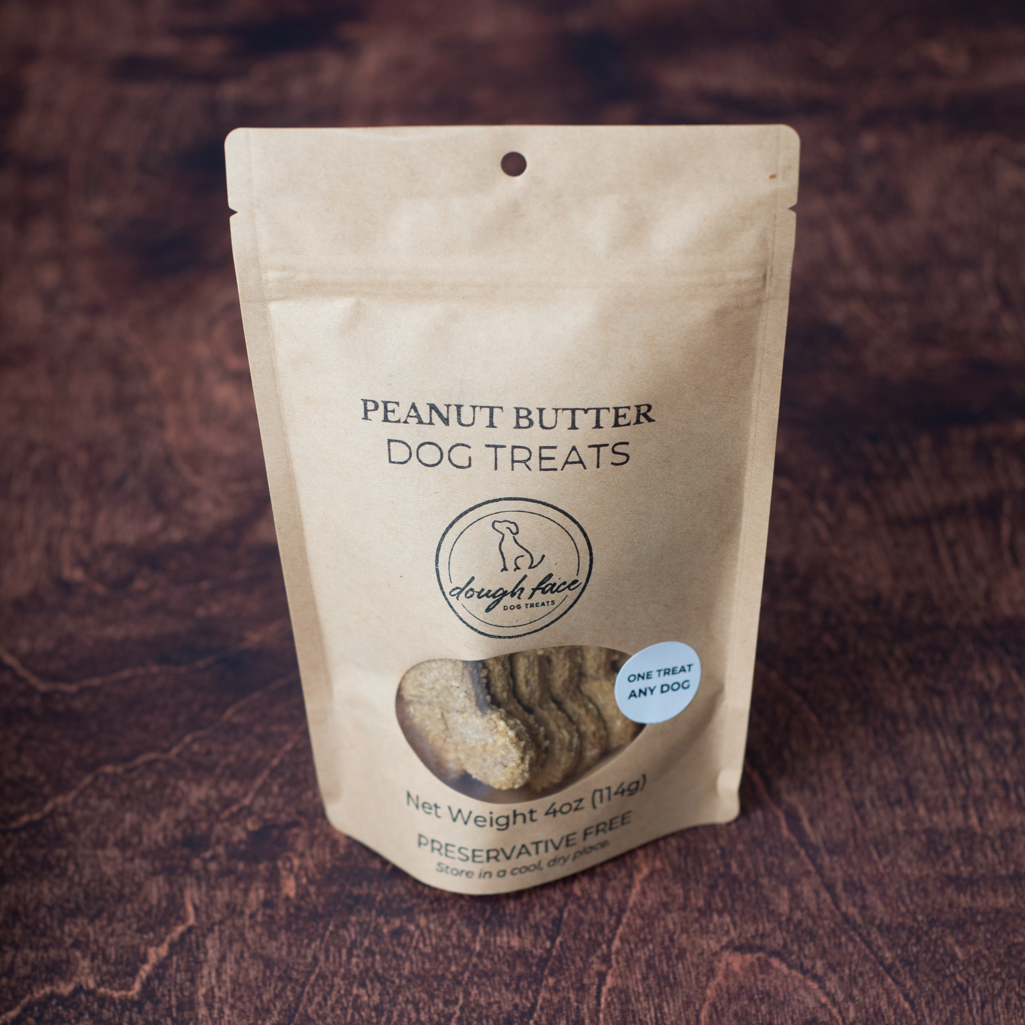 A stand-up pouch of Dough Face Dog Treats Peanut Butter Dog Treats, featuring a clear window that shows a bone-shaped treats inside, set against a dark wood background.