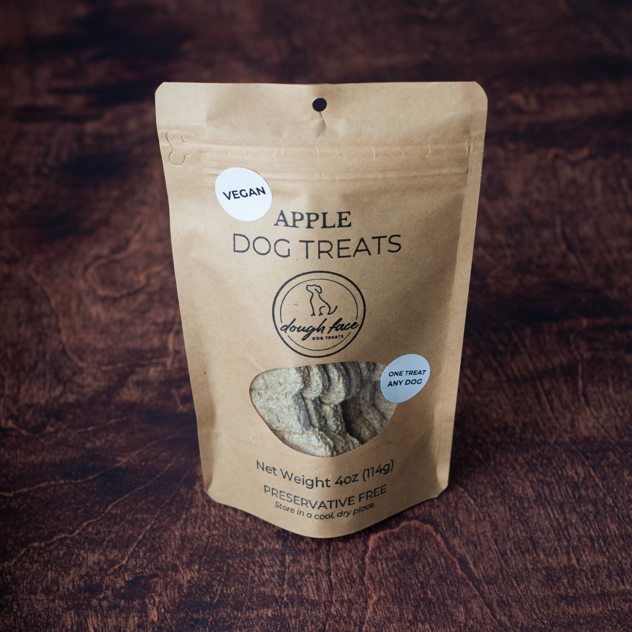 A stand-up kraft paper pouch of Dough Face Dog Treats Apple Dog Treats on a dark brown wood-textured background. The package features a clear window showing the treats inside and displays the Dough Face Dog brand logo.