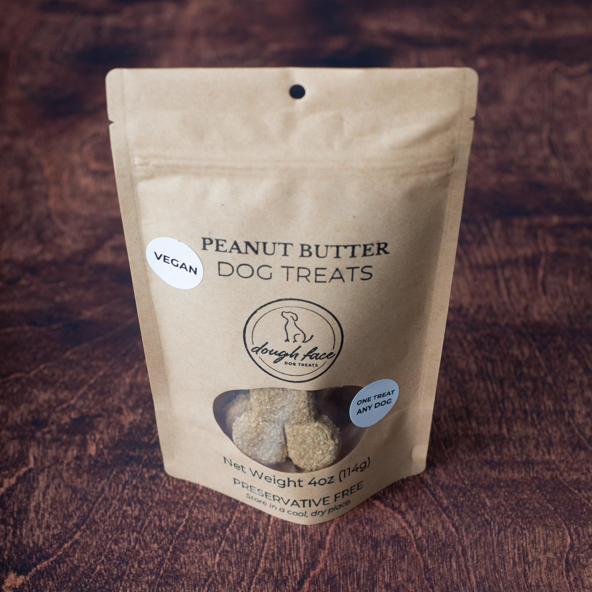 A stand-up pouch of Dough Face Dog Treats Vegan Peanut Butter Dog Treats on a dark wood background. The kraft paper bag features a clear window showing the treats inside and includes a 'VEGAN' badge.