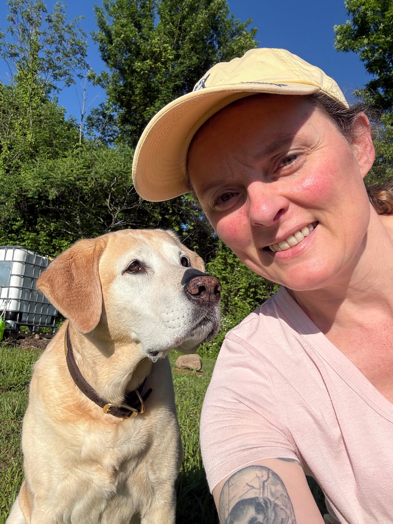 A smiling woman wearing a yellow baseball cap and a light pink t-shirt outdoors next to a white-faced yellow Labrador retriever dog.