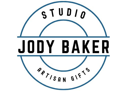 A circular logo with blue outline and black text reading 'STUDIO JODY BAKER ARTISAN GIFTS' on a white background.
