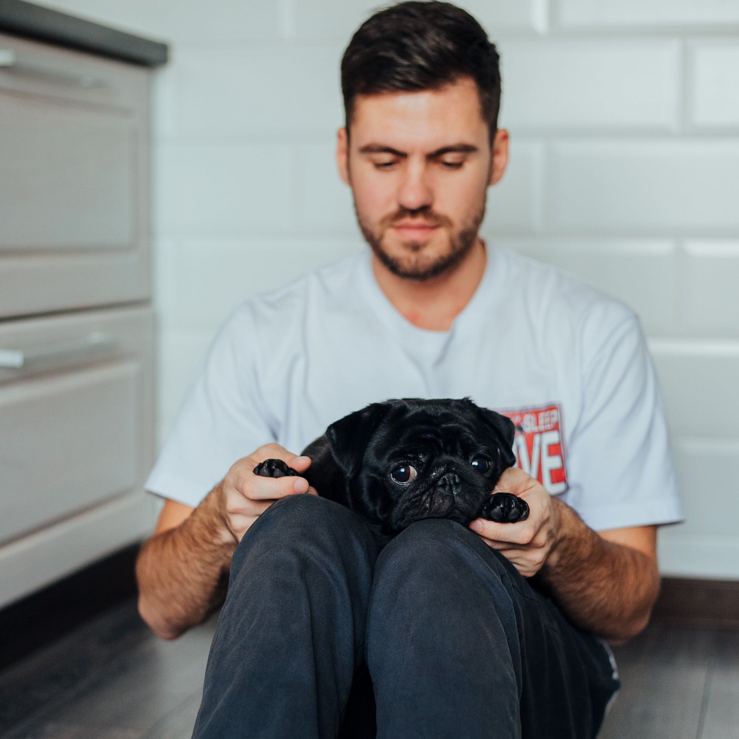 A man sitting on a dark wood floor in a bright room, holding a small black pug dog on his lap. He is wearing a light gray t-shirt and dark gray pants.