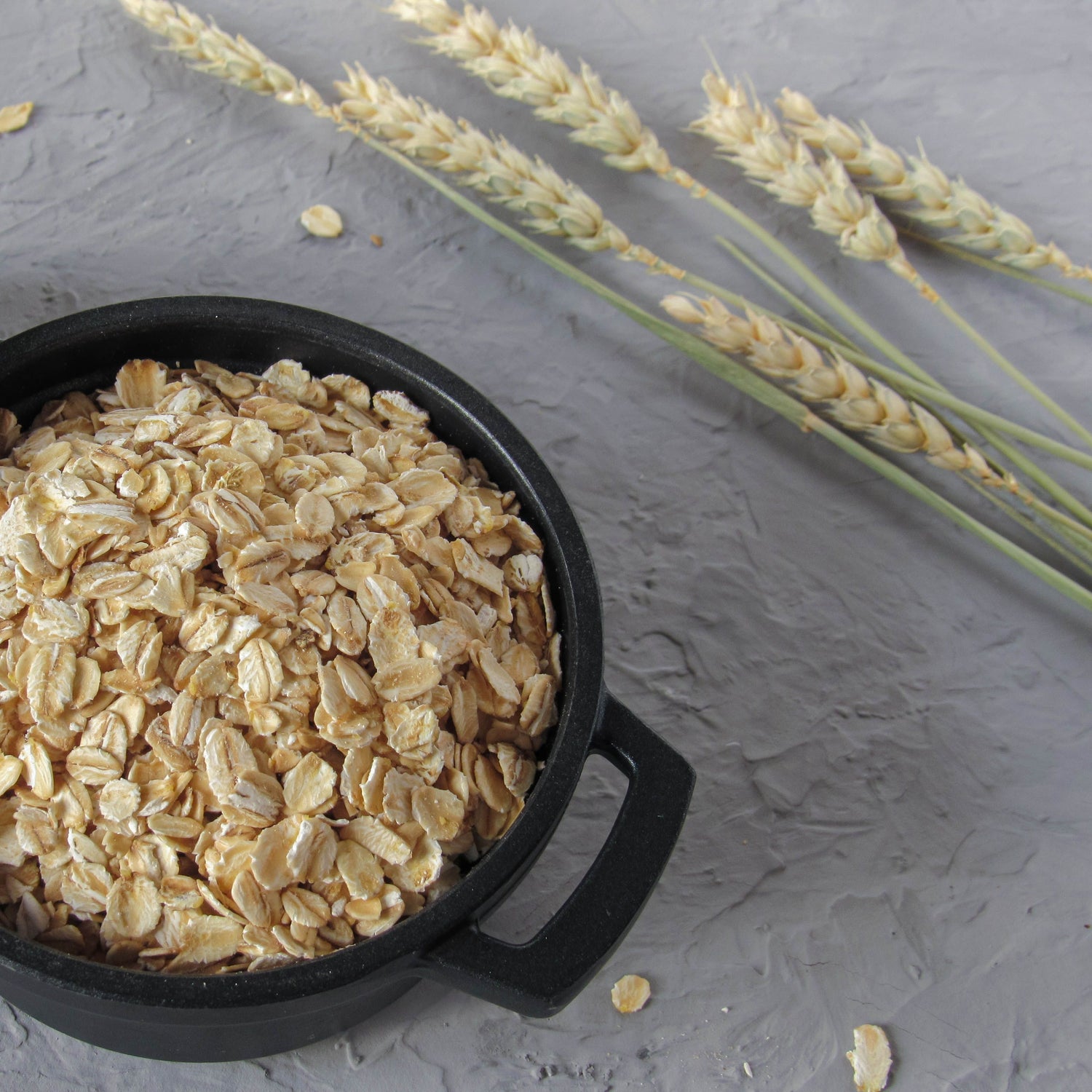 Black pot filled with oats on a gray surface with wheat stalks.