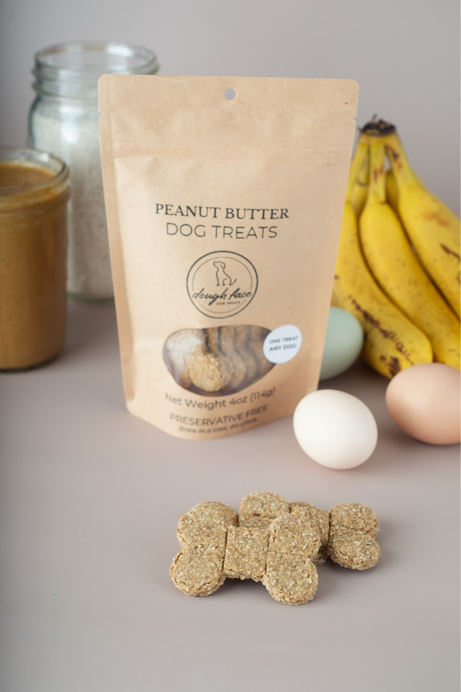 A stand-up pouch of Dough Face Dog Treats' Peanut Butter Dog Treats stands on a gray surface with several bone-shaped treats placed in front. The pouch is beige with black text, and the scene is styled with bananas, eggs, and jars in the background.