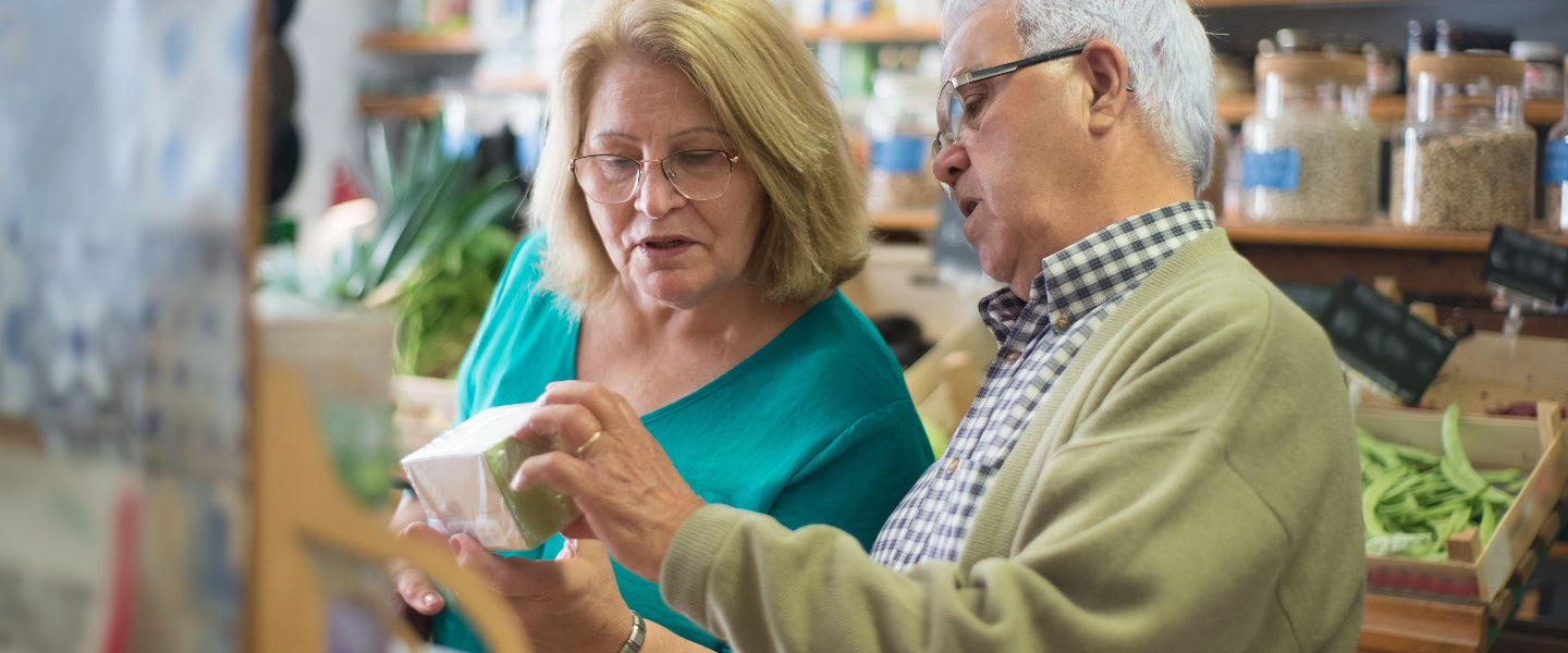 An older man and woman standing in a grocery store aisle, with the man holding a small white paper bag or package. Shelves with colorful produce and jars are visible in the background.