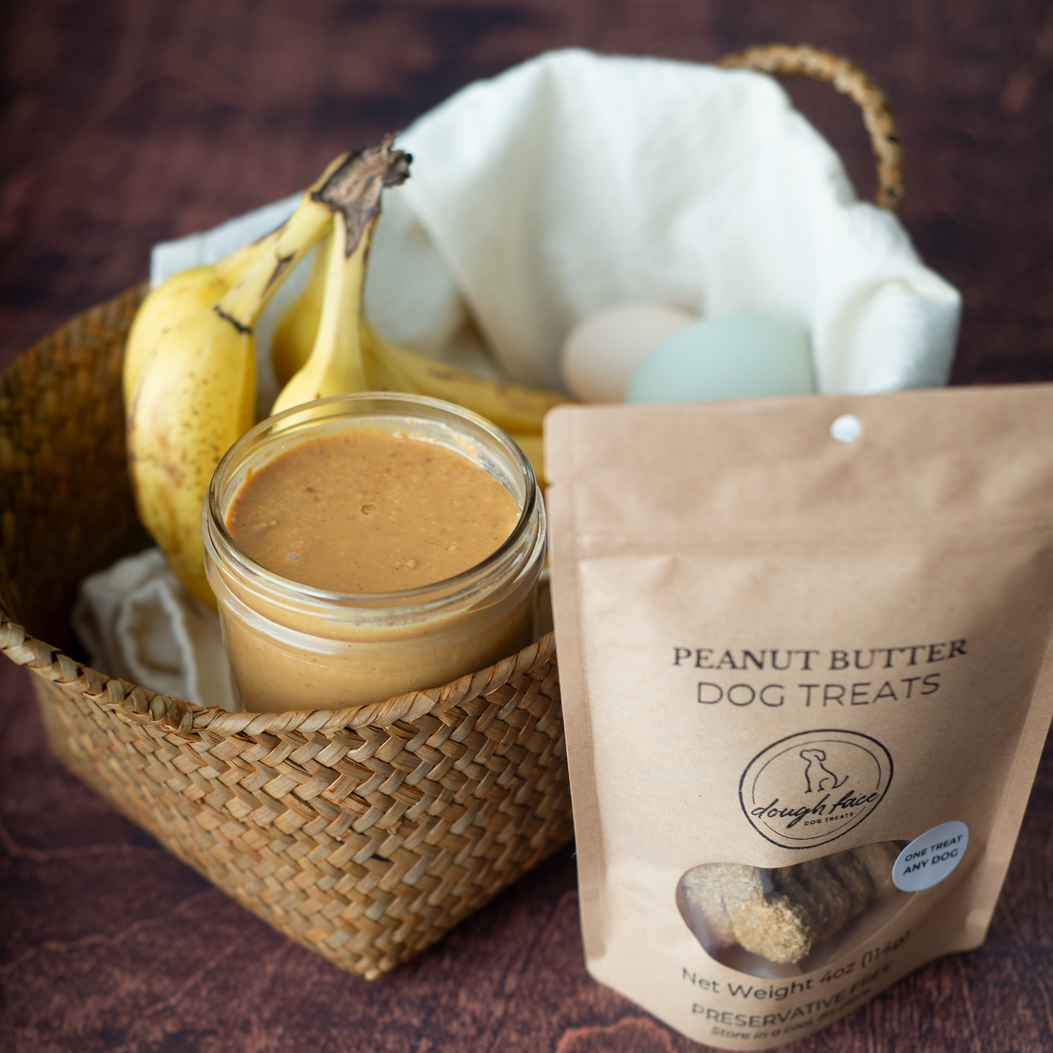 A stand-up pouch labeled 'PEANUT BUTTER DOG TREATS' sits on a dark wooden surface beside a woven basket holding bananas, a jar of peanut butter, and a few dog treats scattered in front. The pouch appears to be the product being sold, while the basket and food props suggest flavor or ingredients.