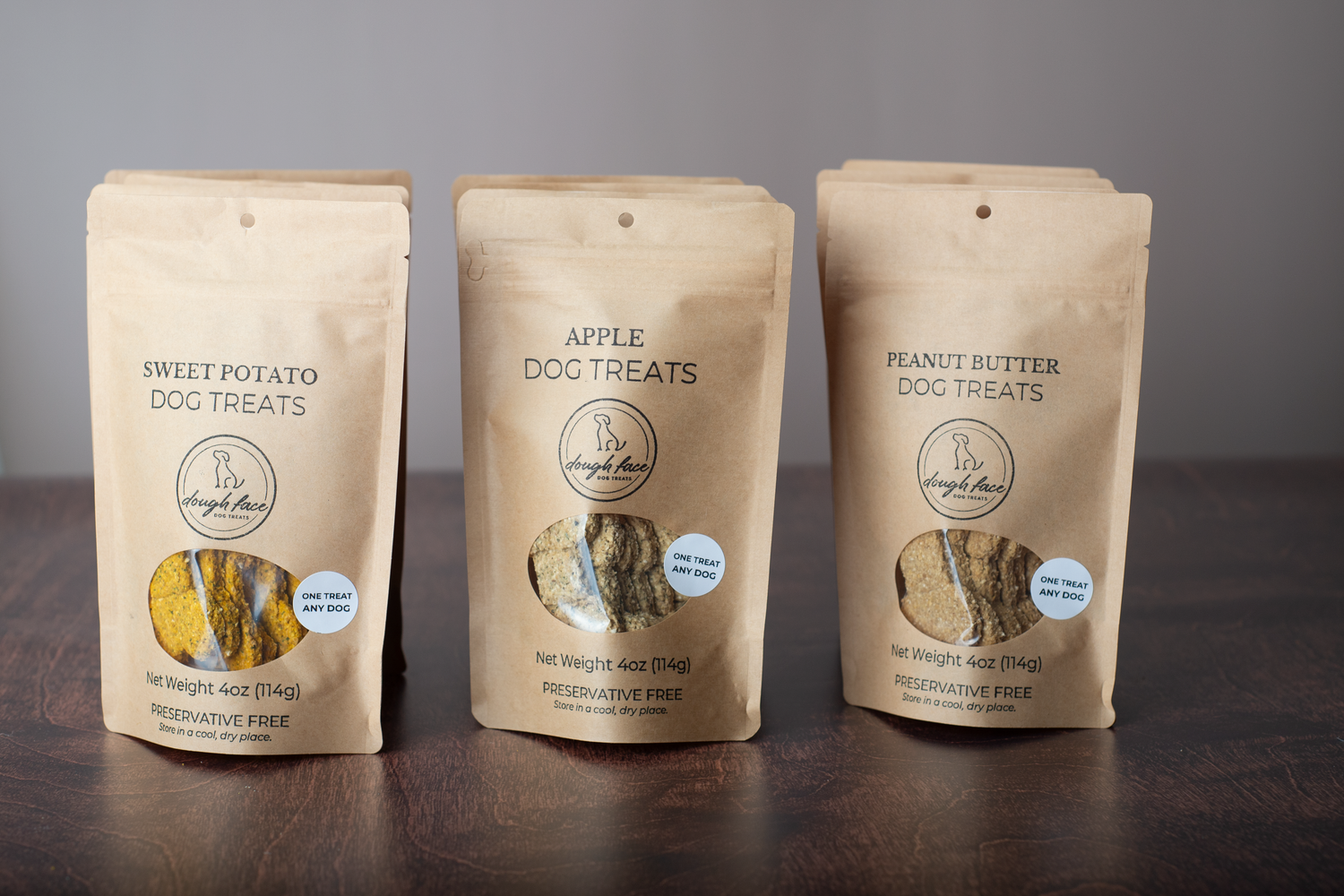 Three stand-up pouches of Dough Face Dog Treats brand dog treats on a dark wooden surface against a gray background. The center pouch is labeled Apple Dog Treats, flanked by Sweet Potato Dog Treats on the left and Peanut Butter Dog Treats on the right.