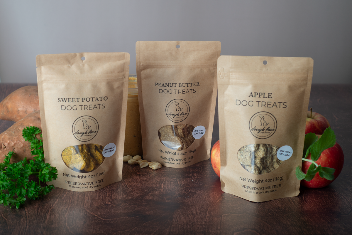 Three stand-up pouches of Dough Face Dog Treats brand dog treats arranged on a dark wooden surface with food props: Sweet Potato Dog Treats on the left, Peanut Butter Dog Treats in the center, and Apple Dog Treats on the right.