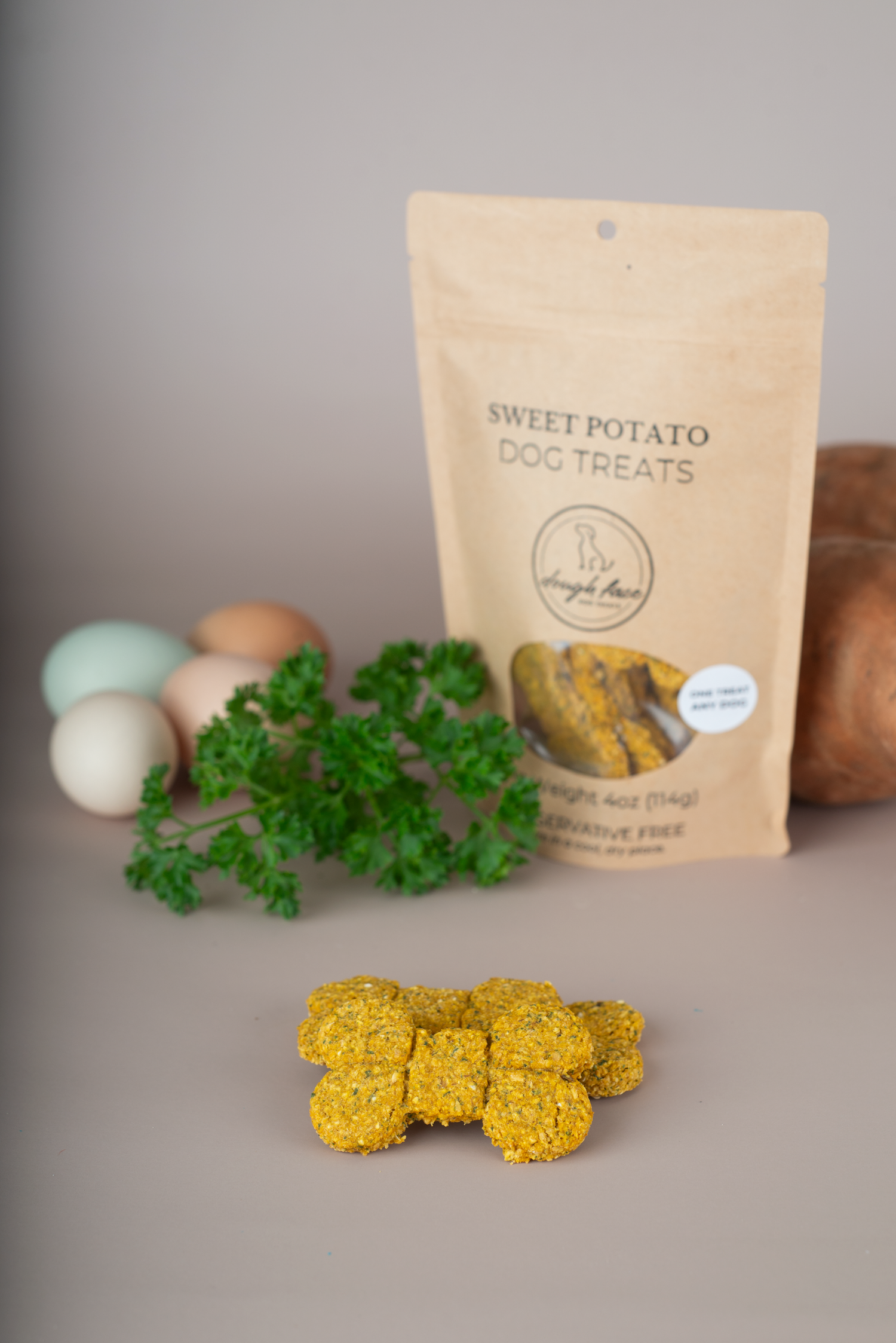 A stand-up pouch of Sweet Potato Dog Treats by Dough Face Dog Treats stands on a neutral gray surface, with several small golden-yellow treat cubes placed in front. Fresh green parsley and a sweet potato are styled around the package, with a soft-focus background