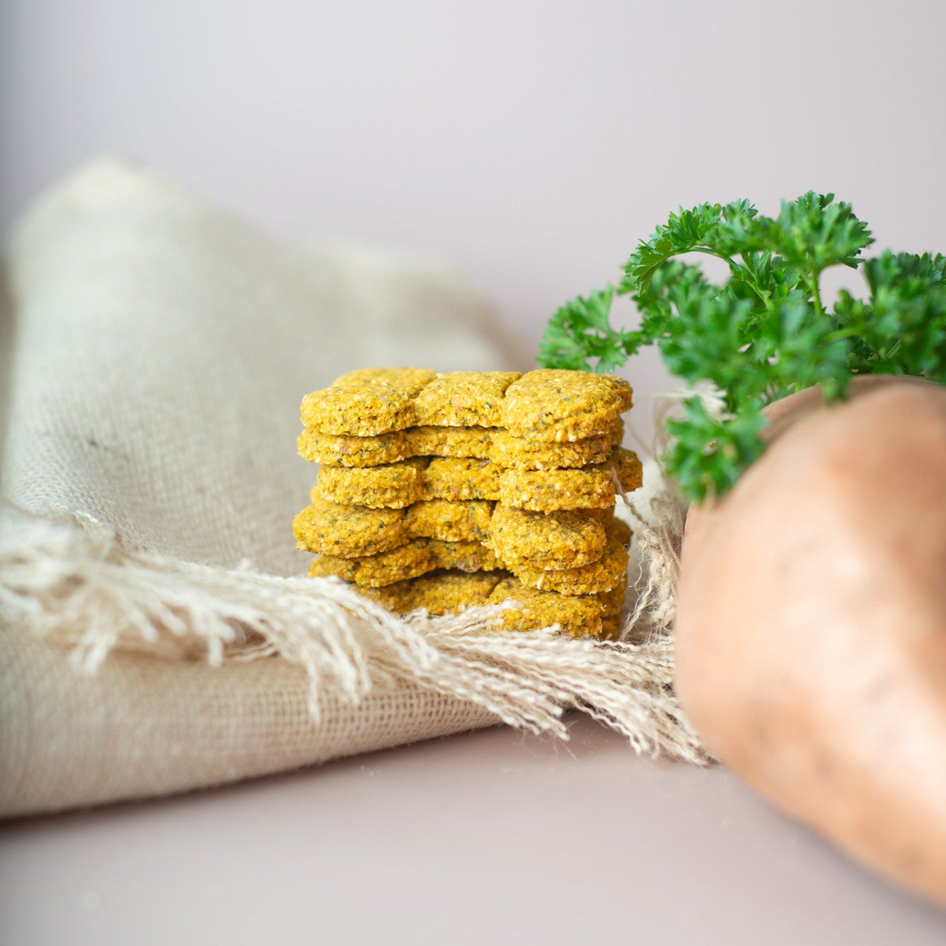 A stack of sweet potato dog treats sits on a beige linen cloth with a sprig of green parsley and a large sweet potato blurred in the foreground, against a soft light gray background.