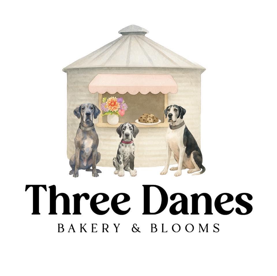 A bakery storefront illustration with three dogs sitting in front of a cream-colored building and a pink awning. Below the illustration is the text 'Three Danes BAKERY & BLOOMS' on a white background.
