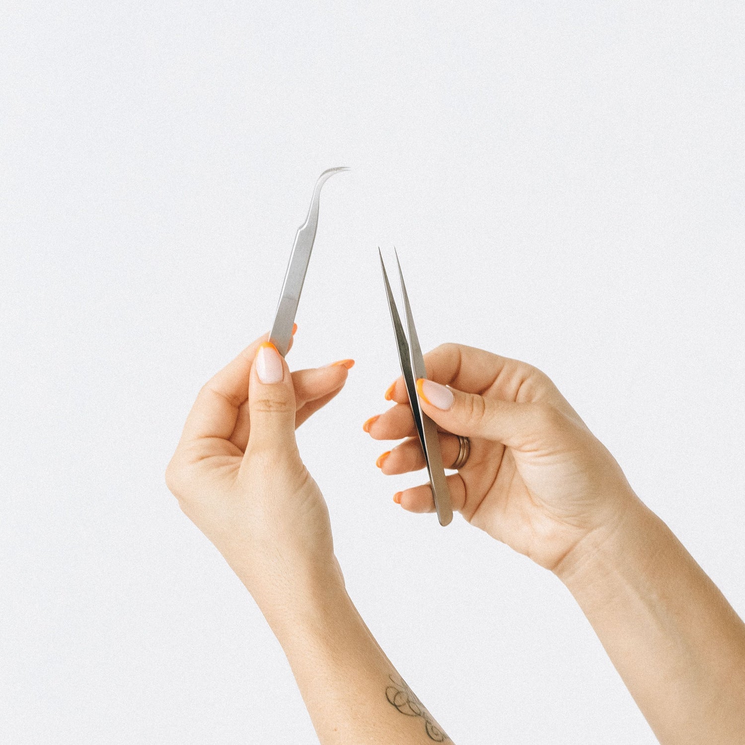 A close-up of a person's hands holding two pairs of silver metal tweezers against a plain light gray background.