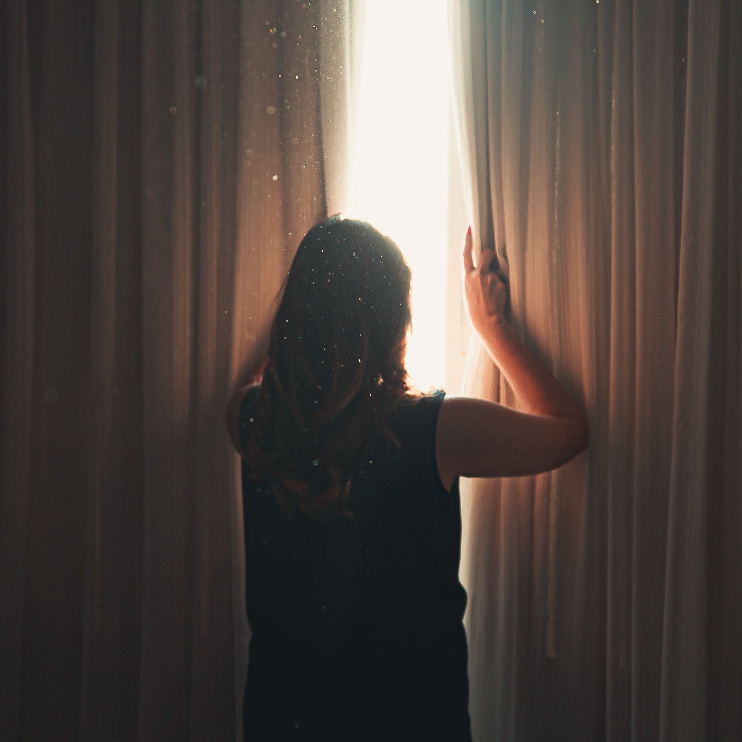 A woman seen from behind standing in front of a bright window framed by sheer curtains, holding the fabric open to let light in. She is wearing a dark sleeveless top, and the scene is backlit with warm golden light.