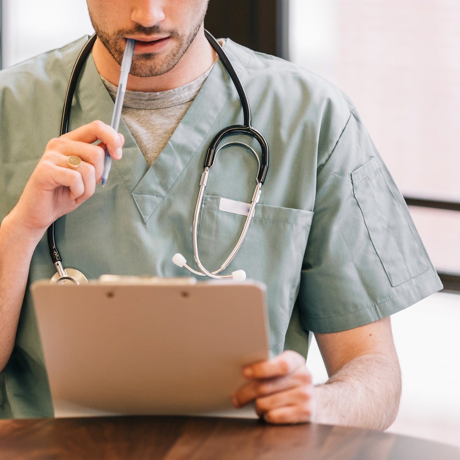 Doctor looking at clipboard with pen in his mouth and a stethoscope around his neck.
